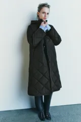 Chaqueta larga acolchada color negro con capucha y manga larga. Tiene bolsillos delanteros y bajo asimétrico con aberturas laterales. El cierre frontal es con botones ocultos por solapa.