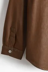 Camisa de corte oversized en denim de algodón rígido revestido, color marrón oscuro. Presenta cuello inglés, tapeta clásica con botones de metal y canesú en la espalda. Tiene hombros caídos, mangas largas con puños abotonados y bajo redondeado.