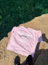Remera rosa de corte recto con estampado en la espalda que incluye el texto "Itta Tours", "Tour", y nombres de ciudades como Ibiza, Madrid, Andorra, Barcelona y Formentera.