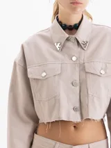 Chaqueta corta estilo jeanero, color beige claro, confeccionada en algodón. Presenta botones metálicos y remaches decorativos en el cuello.
