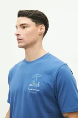 Remera azul de manga corta con cuello redondo y estampado de playa con sombrilla, reposera, balde y pala.