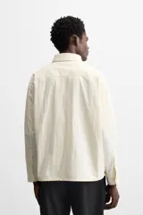 Sobrecamisa color crema de tejido técnico ligero, corte regular fit, cuello solapa, manga larga con puños abotonados, bajo ajustable con elástico en laterales y cierre frontal con cremallera.