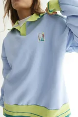 Buzo celeste con cuello polo y puños en rib color verde lima. Tiene un parche con el logo de la marca en el pecho y rayas horizontales en el ruedo.