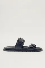 Sandalia plana de cuero negro con dos tiras anchas ajustables mediante hebillas metálicas de estilo envejecido. Presenta una suela gruesa y plana.