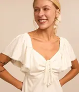 Vestido midi color crudo confeccionado en lino y viscosa, con escote cuadrado, mangas cortas con volados, detalle de lazo en el busto y falda con volados en el ruedo.