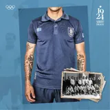 Chomba azul con cuello tipo polo, mangas cortas, escudo de la Asociación Uruguaya de Fútbol en el pecho y bandera de Uruguay en la manga.