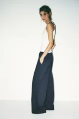 Pantalón negro con finas rayas verticales blancas, de tiro medio y corte ancho. Presenta bolsillos delanteros y traseros, cintura doble tipo bóxer con botones y cierre frontal con cremallera y ganchos metálicos. Detalle de franja marrón en los laterales.
