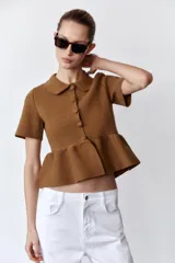 Chaqueta color marrón de tejido de punto, con cuello solapa, manga corta y bajo evasé con volantes. Cierre frontal con botones forrados a tono.