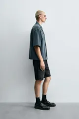 Camisa de denim de algodón, color beige claro, con corte relajado, cuello bowling y manga corta. Presenta cierre frontal con botones a presión y un pequeño bordado a tono en el pecho.