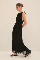 Vestido largo tipo musculosa con escote profundo en el frente. Confeccionado en gasa con forrería a tono. Posee corte en la cintura acompañado de un cinturón de la misma materialidad. Tiene un volado en gasa que se extiende por toda la abertura del cuello. Posee lazos en el cuello, que permiten atar la prenda con plumas metálicas en su terminación.