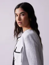 Chaqueta de tweed blanca con ribete negro, escote a la base, abertura central con botones, bolsillos delanteros y puños con aplique de botones perlados.