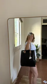 Bolso tote negro de cuero sintético con textura de cocodrilo. Presenta costuras decorativas en los laterales y un dije colgante en forma de cruz plateada con detalles de perlas.