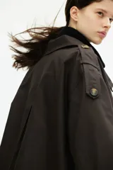 Gabardina corta color marrón chocolate, de corte holgado, con cuello clásico, doble botonadura, charreteras, trabillas abotonadas en los puños, bolsillos delanteros con solapa y pliegue invertido en la espalda. Forrada.