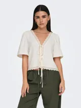 Top blanco con textura, escote en V, mangas cortas abullonadas y cierre con lazos. Detalle de encaje en escote y ruedo.