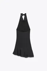 Vestido corto negro de tejido fluido con cuello halter que se ata con lazada. Tiene forro interior, detalle de frunces y bajo asimétrico con volantes. Cierre lateral con cremallera oculta.