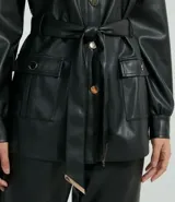 Chaqueta larga de cuero sintético negro, con cuello camisero, cierre frontal con botones, cinturón con hebilla, bolsillos con solapa y puños abotonados.