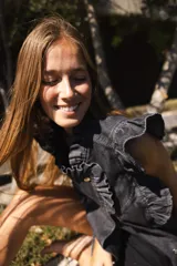 Chaleco de denim color topo o gris claro lavado, con volados estructurados en los hombros y a lo largo del frente. Es de corte recto, sin mangas, con cierre frontal de botones metálicos color cobre y bolsillos laterales.