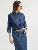 Camisa celeste de denim suave, corte relajado, con cuello clásico, cierre frontal con botones marrones, manga larga con puños abotonados, dos bolsillos de parche en el pecho y ruedo redondeado.