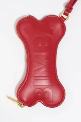 Monedero pequeño con forma de hueso, color gris claro, con cierre de cremallera dorada y correa de mano a juego. Presenta el logo de la marca estampado en dorado en el centro.