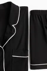 Pijama corto de dos piezas color negro, en punto suave con ribetes blancos en contraste. Camisa de manga corta con cuello abierto, botones al frente y bolsillo superior abierto. Short con elástico en la cintura.