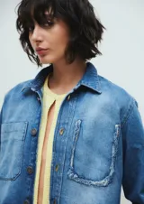 Campera corta de denim azul con efecto desgastado. Tiene cuello clásico, cierre frontal con botones, dos bolsillos delanteros y terminaciones desflecadas.