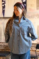 Camisa de rayas verticales azules y blancas, con cuello camisero, manga larga y puños abotonados. Presenta un bolsillo en el pecho con ribete rojo y un detalle de ribete rojo en el cuello y la tapeta de botones.