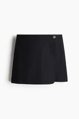 Short pollera negro de tiro alto, cruzado y con botón a un lado. Confeccionado en sarga de algodón, con cierre oculto adelante y bolsillos insertados decorativos atrás.