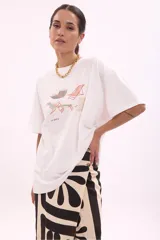 Remera blanca de algodón, corte oversize, con cuello redondo y manga corta. Presenta un estampado frontal con varias reposeras y hamacas de playa, y el texto "LA SIESTA".