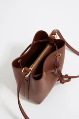 Bolso tipo bucket de cuero color marrón con cierre de cordón ajustable. Tiene correa de hombro delgada y logo de la marca estampado en dorado en el frente.