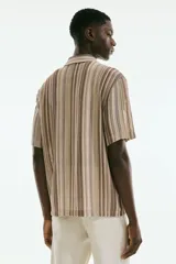 Camisa de manga corta con cuello inglés, botones adelante y bajo recto. Tejido de punto pointelle con estilo crochet y diseño a rayas verticales en tonos beige, marrón y blanco.