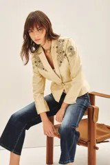 Saco sastre color beige claro, de manga larga y calce al cuerpo, con cierre frontal ajustable mediante cinturón con hebilla de piedras. Presenta bordados florales en tonos dorados y negros con lentejuelas y canutillos en los hombros y solapas.