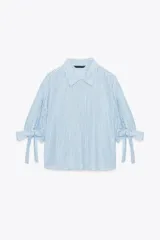 Camisa celeste y blanca a rayas verticales, confeccionada con lino e hilo metalizado. Tiene cuello solapa, mangas 3/4 con lazos en los puños y cierre frontal con botones ocultos por solapa.