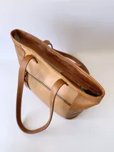 Bolso tipo tote de cuero color camel con paneles laterales texturizados. Tiene doble asa de hombro, cierre superior con cremallera y bolsillos internos.