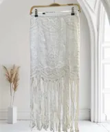 Pollera blanca de largo midi, con intrincado bordado de encaje floral y detalles calados. Presenta un borde festoneado con flecos largos y anchos que caen desde el ruedo.