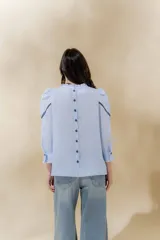 Blusa de lino blanca con escote a la base y terminación de voladito al bies desflecado. Delantera con alforzas verticales y mangas románticas tres cuartos, con detalle de puntilla. Puños con alforzas y voladitos que suman un toque femenino. Espalda abotonada.