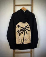 Buzo hoodie negro de corte oversize con capucha y cordón ajustable. Estampado en la espalda con diseño de dos palmeras y el sol dentro de un arco.