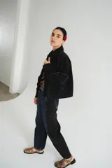 Campera de jean negra tipo bomber con cuello redondo y cierre frontal con botones metálicos. Presenta un parche rectangular con diseño gráfico en la espalda.