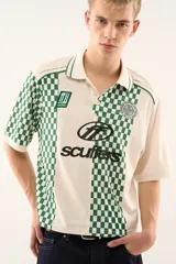 Polo de manga corta color blanco crudo con cuello y ribetes verdes. Presenta un diseño inspirado en camisetas de fútbol con bloques de cuadros verdes y blancos en las mangas y laterales, y una franja central blanca con estampado de logo en negro.
