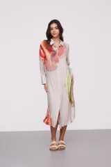Vestido camisero midi color beige con estampado abstracto en tonos naranja, rojo, verde y amarillo. Tiene cuello camisero, mangas largas con puños abotonados y cierre frontal con botones.