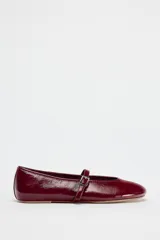 Zapato tipo bailarina color borgoña con efecto acharolado, acabado en punta redonda y tira con hebilla ajustable en la parte delantera.