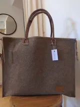 Bolso grande tipo tote de fieltro color marrón con asas de cuero marrón. Tiene un parche rectangular de cuero marrón en la parte inferior frontal y un tag colgante con el nombre de la marca.