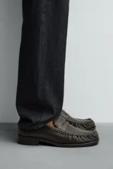 Mocasín de cuero color marrón oscuro con detalle de antifaz en el empeine, plantilla de cuero, horma cuadrada y suela a tono con ligero tacón.