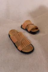 Sandalias bajas de cuero color marrón claro, con suela de goma negra y detalle de flecos en la tira delantera.