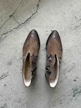 Bota corta de cuero color marrón, con taco cuadrado y punta fina. Presenta una correa con tachas pequeñas y una argolla metálica decorativa alrededor del tobillo. Cuenta con cierre lateral.