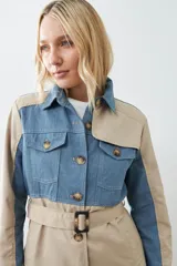 Trench largo color beige con secciones en denim azul, cuello camisero, cierre frontal con botones y bolsillos con solapa en el pecho.
