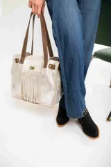 Bolso tipo tote de cuero color plateado con flecos en la parte frontal, doble asa de hombro marrón y cierre con broche metálico.