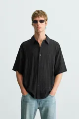 Camisa de hombre de manga corta, cuello solapa y corte relaxed fit. Tejido blanco con textura sutilmente arrugada o plisada. Cierre frontal de botonadura y bajo con aberturas laterales.