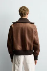 Cazadora de cuero color marrón, estilo bomber, con cuello ancho de punto acanalado marrón oscuro, cierre frontal de cremallera y puños y cintura elásticos de punto acanalado marrón oscuro.