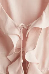 Blusa color rosa pálido de lino y viscosa, con escote en V pronunciado, mangas cortas y volados en el escote, mangas y ruedo. Se ata en el centro con dos cordones finos.
