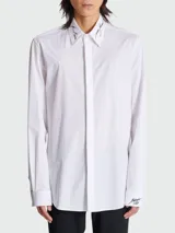 Camisa blanca de popelina de algodón con cuello clásico, cierre frontal con botones ocultos, mangas largas con puños abotonados y bordado con el logo de la marca en cuello y puños.